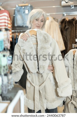 Customer in store has removed hanger with fur coat from window and is considering product. Senior woman represents comfort and warmth that this coat will give on cold winter evenings
