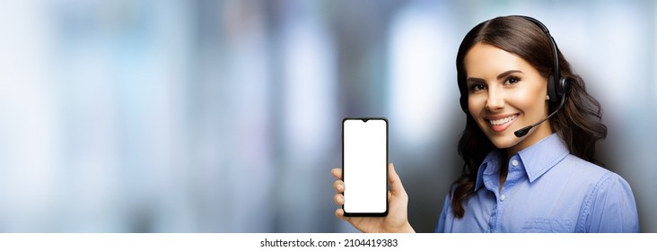 Customer support phone operator in headset holding showing smartphone cell phone mobile with white blank mock up screen blurred office interior background. Consulting and assistance service callcenter