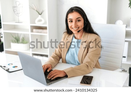 Customer support service. Arabian or indian beautiful woman, call center operator with headset, working on support hotline in a modern office with a laptop, looking at the camera, smiling friendly