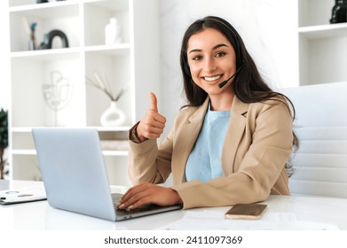 Customer support service. Confident positive woman, call center operator with headset, working on support hotline in a modern office with a laptop, looks at the camera, smiling, shows thumb-up gesture - Powered by Shutterstock