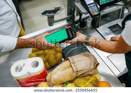 Customer using smartphone with green screen for contactless payment at supermarket checkout, with cashier holding payment terminal and groceries on conveyor belt