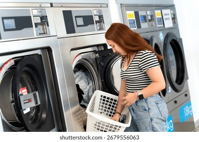 Customer using a washing machine in a self-service laundry, putting clothes inside while holding a white basket