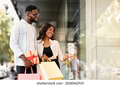 Customers And Consumerism Concept. Smiling African American couple looking and pointing at mall window, walking near shopping mall. Seasonal Sales And Discount, Purchase And Retail