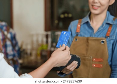 Customer's hand presenting a blue credit card to a barista at a coffee shop register. Focus on card and terminal, with the smiling barista in the background. Cashless payments. - Powered by Shutterstock