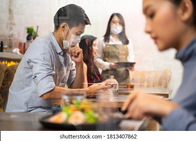 Customers In New Normal Restaurant Sitting At Seperate Desk With Glass  Partition In Between. Mask Worn By Customer Waiting For Food Playing With Mobile Phone And Waitress Taking Orders Using Ipad. 