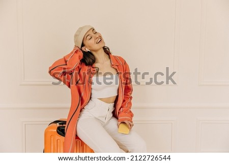Cut beauty lady sits on suitcase and listens to music headphones through phone white background. Young brown-haired woman enjoys new song with eyes closed. Audio technology, and music concept.