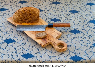 Cut bread loaf on wooden olive wood board. Knife made from modern Damascus steel made by Japanese artisan. Table cloth on table.