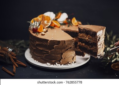 A Cut Of Chocolate Cake On A Wooden Dark Background Adorned With Citrus And Rosemary. The Confectioner's Hand Holds A Piece Of Cake On The Patisserie.