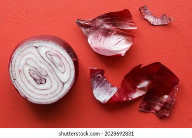 A cut half of a red onion lies on a red background next to a peeled onion husk. Close-up