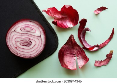 A cut half of a red onion lies on a black and light green background next to a peeled onion husk. Close-up