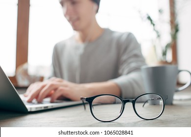 Cut view of a guy sitting at the table and wokring. He is wokring using the laptop. Besides the laptop there is a cup of tea. There are glasses in the front of the picture