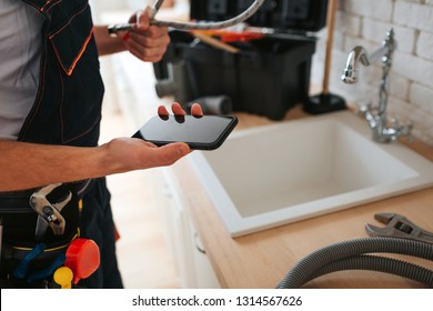 Cut View Of Man Standing In Kitchen At Sink. He Hold Phone And Wrench. Hose On Desk.