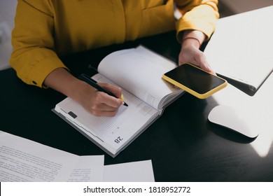 Cut View Of Woman's Hands. Caucasian Woman In Yellow Blouse Writing In Diary Or Planner And Looking At Phone. Top View. Papers On Black Desk. Red Bracelet On Hand. Work At Home.