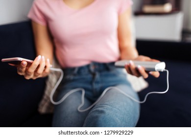 Cut view of young woman hold phone with charge plugged in to it. She hold them in both hands. Model sit on sofa in room.