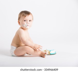 a cute 1 year old sits in a white studio setting. The boy looks to his left with a face full of cake frosting with half eaten cake. He is only dressed in a white diaper