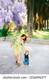 Cute 18 months old baby is walking hand-in-hand with his mother under a wysteria tree