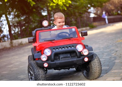 Cute 2 Year Old Boy In A White T Shirt Is Riding A Red Electric Car In The Park