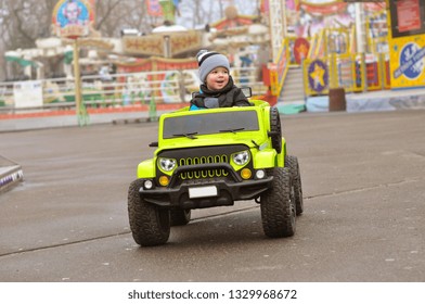 Cute 2 Year Old Boy In A Hat And Jacket Is Riding A Green Electric Car In The Amusement Park.