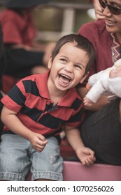 Cute 2 Year Old Mixed Race Asian Caucasian Boy Laughing And Playing With His Mom And Doll Sitting At A Sporting Event