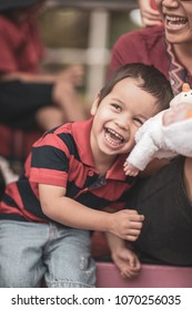 Cute 2 Year Old Mixed Race Asian Caucasian Boy Laughing And Playing With His Mom And Doll Sitting At A Sporting Event