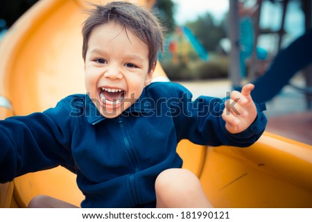 Cute 3 year old boy excitedly plays on a yellow playground slide on a cool cloudy day