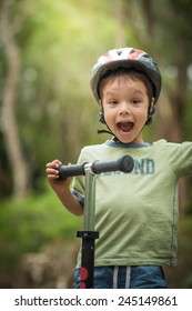 Cute 4 Year Old Mixed Race Asian Caucasian Boy Wearing A Bike Helmet Cheerfully Plays On His Scooter On A Green Forest Road