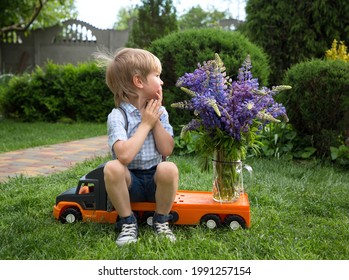 cute 4-5 year old boy in shorts and shirt sits on toy orange truck with bouquet of lupine flowers. little courier, gift to friend, with love for mom. grandmother. Flower delivery concept. Positive