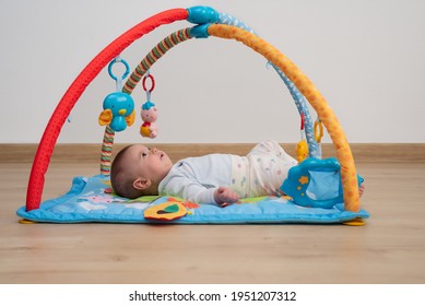 Cute 5 Months Baby Girl Lying Face Up On A Blue Play Mat While Is Looking At Toys Of A Baby Gym.   Games For Psychomotor Development. 