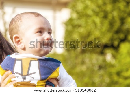 Cute 6 months old baby with Light brown hair in white, blue and brownish long-sleeved shirt is embraced and held by his mother: he is biting his lips and puffing his cheecks in a funny face