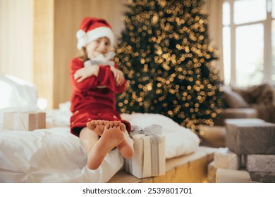 Cute 7-years old girl wearing Santa hat and holiday pajamas enjoying her a Christmas presents in a cozy festive bedroom.