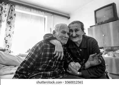 Cute 80 Plus Year Old Senior Married Couple Hugging And Smiling Portrait. Black And White Waist Up Image Of Happy Elderly Couple Sitting On A Bed In A Nursing Home.