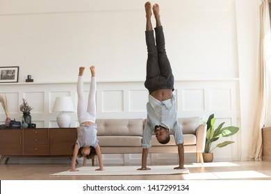 Cute active african kid girl copy imitate father doing gymnastic handstand exercise in living room, sporty family black dad and child daughter stand on hands upside down having fun together at home