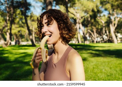 Cute Active Redhead Woman Wearing Sportive Clothes Relaxing After A Long Workout In The City Park And Eating A Banana. Healthy Food, Outdoor Sport Concepts.