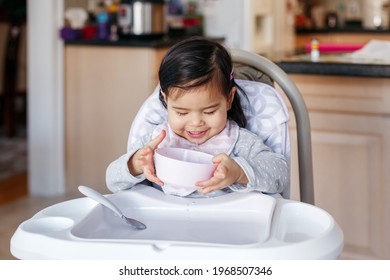 Cute Adorable Asian Chinese Kid Girl Sitting In High Chair Eating Soup With Spoon. Healthy Eating For Kids Children. Toddler Eating Independently In Kitchen At Home. Candid Real Authentic Moments.