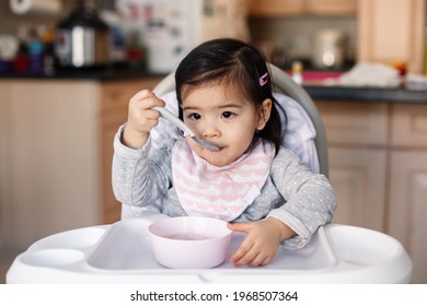 Cute Adorable Asian Chinese Kid Girl Sitting In High Chair Eating Soup With Spoon. Healthy Eating For Kids Children. Toddler Eating Independently In Kitchen At Home. Candid Real Authentic Moments.