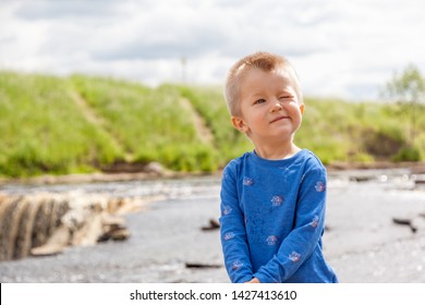 Cute Adorable Baby Boy Is Blinking, Closing His Eye From Sunshine, Grimacing On Sightseeng Excursion On River Waterfall On Summer Day