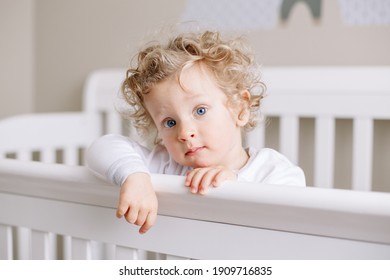 Cute adorable baby boy toddler standing in crib at kids nursery room at home. Curious charming baby boy with curly blond hair and blue eyes looking at camera. Happy authentic candid home life.  - Powered by Shutterstock