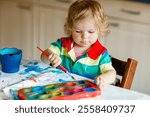 Cute adorable baby girl learning painting with water colors. Little toddler child drawing at home, using colorful brushes. Healthy happy daughter experimenting with colors, water at home or nursery.