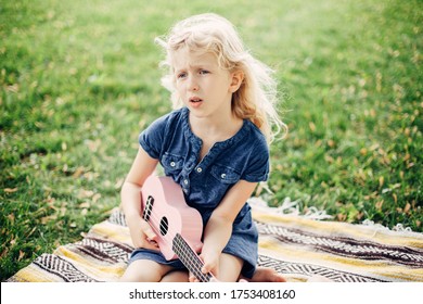 Cute Adorable Blonde Girl Playing Pink Guitar Toy Outdoor. Child Playing Music And Singing Song In Park. Hobby Activity For Children Kids. Tender Memorable Authentic Candid Childhood Moment. 