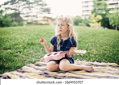 Cute Adorable Blonde Girl Playing Pink Guitar Toy Outdoor. Child Playing Music And Singing Song In Park. Music Hobby Activity For Children Kids. Tender Memorable Authentic Candid Childhood Moment. 