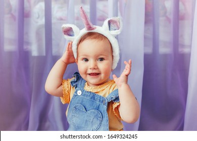 Cute adorable Caucasian baby girl wearing unicorn headband with horn and ears. Pretty funny child kid with head hair fairy tale animal accessory toy. Smiling toddler playing at home looking at camera.