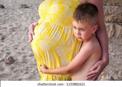 Cute Adorable Caucasian Child With An Anxious Look Embracing His Pregnant Mother's Belly. Expecting A Child, Expecting A Brother, Expecting A Sister. Newborn Brother Sister Jealousy, Jealous Child