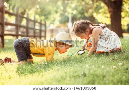 Cute adorable Caucasian girl and boy looking at plants grass in park through magnifying glass. Children friends siblings with loupe studying learning nature outside. Child education concept.