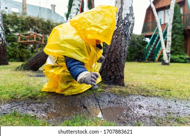 Cute Adorable Caucasian Toddler Boy In Bright Yellow Raincoat And Wellies Playing Alone At Dirt Muddy Puddle During Cold Spring Rainy Day At Home Backyard. Carefree Childhood Concept. Bad Weather