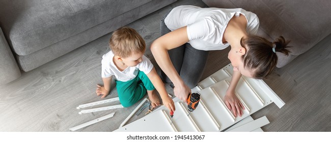 Cute adorable caucasian toddler boy kid sit on floor and help mom assembling furniture shelf with power screwdriver tool. Young adult mum with funny little child enjoy playing at home indoors