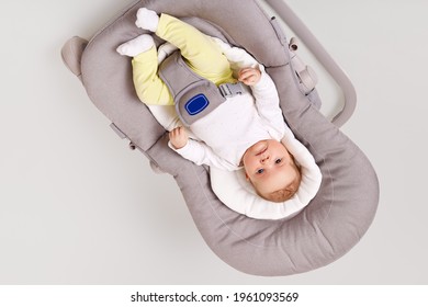 Cute adorable child wearing white shirt and yellow trousers posing in gray bouncer chair, looking around her and exploring world, isolated over light background.