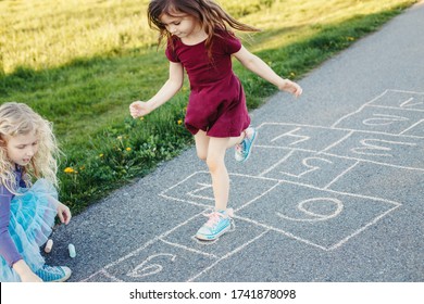 Cute Adorable Children Girls Friends Playing Jumping Hopscotch Outdoor. Funny Activity Game For Kids On Playground Outside. Summer Backyard Street Sport For Children. Happy Childhood Lifestyle.