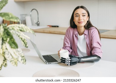 Cute adorable female of 20s with long lashes and sensual lips sitting at table with white cup of coffee in hands, one arm made of metal bionic mechanical prosthesis after amputation