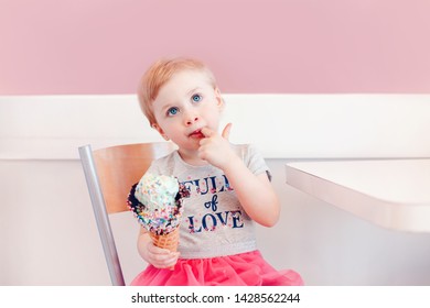 Cute Adorable Funny Caucasian Blonde Babyl Girl Child With Blue Eyes Eating Ice Cream In Large Waffle Cone With Colorful Sprinkles. Kid Licking Fingers. Tasty Summer Food And Happy Childhood