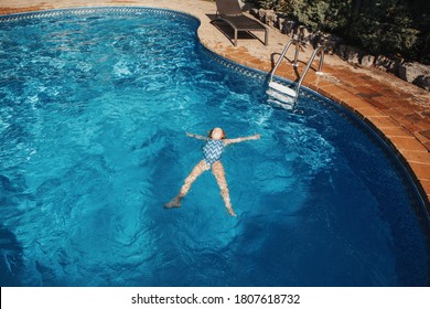 Cute Adorable Girl Floating On Back In Water Pool On Backyard. Kid Child Enjoying Having Fun In Swimming Pool. Summer Outdoor Water Activity For Kids. View From Above Overhead.